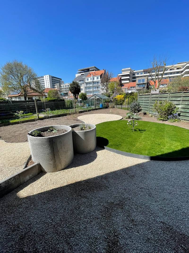 Rond gazon tussen lichte grindvlakken, met twee ronde betonnen plantenbakken op de voorgrond
