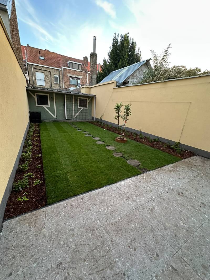 Dezelfde stadstuin na de aanleg, met nieuw gazon, stapstenen en borders met houtsnippers tussen gele tuinmuren